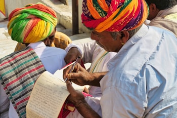 A group of people wearing colorful traditional attire, with one person in a vibrant turban writing or reviewing a paper using a pen. Another person also dressed in traditional clothing holds a notepad, which appears to have handwritten notes. The setting is outdoors with natural lighting.