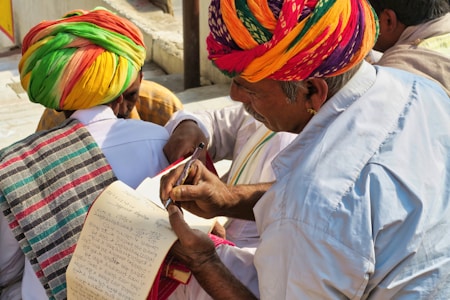 A group of people wearing colorful traditional attire, with one person in a vibrant turban writing or reviewing a paper using a pen. Another person also dressed in traditional clothing holds a notepad, which appears to have handwritten notes. The setting is outdoors with natural lighting.