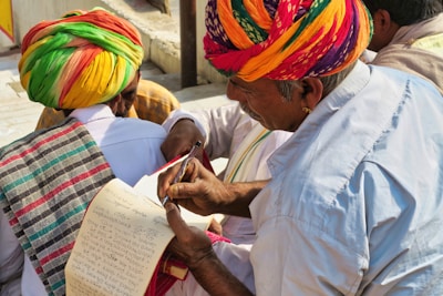 A group of people wearing colorful traditional attire, with one person in a vibrant turban writing or reviewing a paper using a pen. Another person also dressed in traditional clothing holds a notepad, which appears to have handwritten notes. The setting is outdoors with natural lighting.
