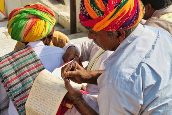 A group of people wearing colorful traditional attire, with one person in a vibrant turban writing or reviewing a paper using a pen. Another person also dressed in traditional clothing holds a notepad, which appears to have handwritten notes. The setting is outdoors with natural lighting.