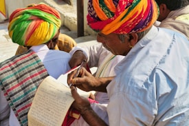 A group of people wearing colorful traditional attire, with one person in a vibrant turban writing or reviewing a paper using a pen. Another person also dressed in traditional clothing holds a notepad, which appears to have handwritten notes. The setting is outdoors with natural lighting.