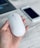 A white, sleek computer mouse is held in a person's hand, with a laptop and a smartphone in the background on a wooden surface.