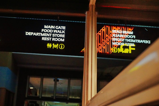 A sign displaying directions to various locations like the main gate, food walk, department store, and restroom is visible. The text is illuminated in white and surrounded by icons for facilities. The reflection of these words and symbols appears on a glass surface nearby, showing some red decorative patterns.