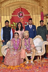 A wedding scene with a bride and groom seated on a decorative couch, adorned with traditional Indian garments. The bride wears a red and gold lehenga, while the groom is in a sherwani. Both have floral garlands around their necks. Standing behind them are four individuals dressed in formal attire. The background features ornate gold and red decorations with floral arrangements.