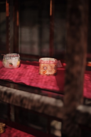 Two decorative ceramic cups are placed on a red surface, one with a plaid and strawberry pattern and the other with a pink design featuring circular motifs. The scene is softly lit, casting shadows on the surface, creating a warm and intimate atmosphere.