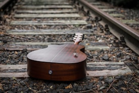 A guitar lies face-down on a set of train tracks. The wood of the railway ties and the gravel fill in the spaces between the tracks. Dry leaves and twigs are scattered around, adding to the overall rustic and slightly abandoned feel of the scene.