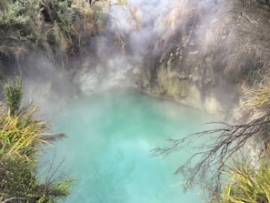 A serene geothermal pool surrounded by rocky cliffs and lush vegetation. Mist rises from the turquoise water, creating an ethereal and mystical atmosphere. The foliage includes a mix of green shrubs and grasses, adding to the natural and tranquil setting.
