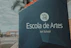 A large signboard with the text 'Escola de Artes Art School' in both Portuguese and English stands prominently by the roadside. The background features palm trees and a few buildings with a cloudy sky.