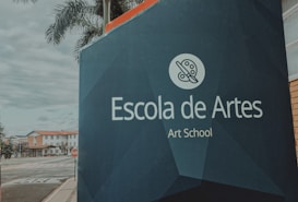 A large signboard with the text 'Escola de Artes Art School' in both Portuguese and English stands prominently by the roadside. The background features palm trees and a few buildings with a cloudy sky.