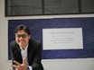 a man in a suit and glasses sitting in front of a sign
