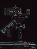 A high-tech gimbal stabilizer supports a smartphone displaying a camera interface. The setup is surrounded by a dark background, highlighting the device's buttons and branding with red accents.
