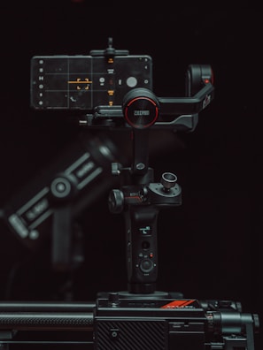 A high-tech gimbal stabilizer supports a smartphone displaying a camera interface. The setup is surrounded by a dark background, highlighting the device's buttons and branding with red accents.