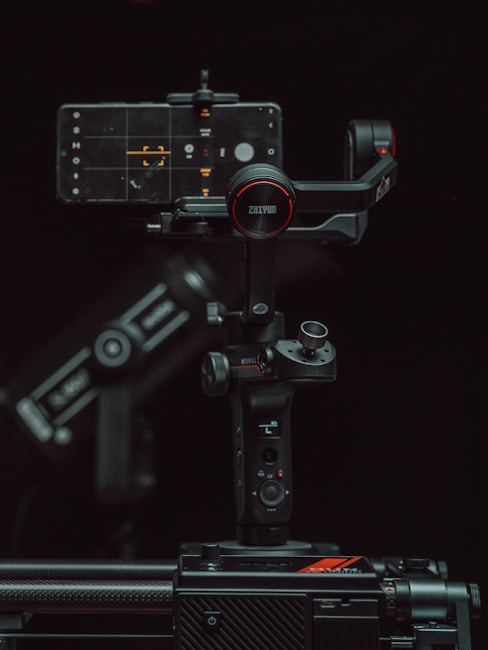 A high-tech gimbal stabilizer supports a smartphone displaying a camera interface. The setup is surrounded by a dark background, highlighting the device's buttons and branding with red accents.