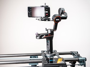 A smartphone is mounted on a motorized gimbal which is attached to a slider. The setup is designed for filming steady video shots. The gimbal has a sleek black design with red accents and features multiple control buttons and adjustment knobs. The background is plain and well-lit, drawing focus to the equipment.
