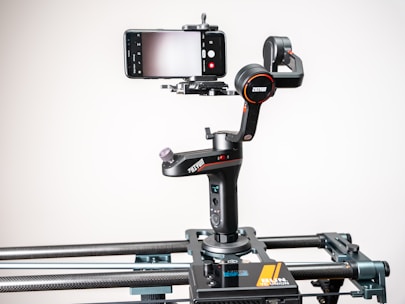A smartphone is mounted on a motorized gimbal which is attached to a slider. The setup is designed for filming steady video shots. The gimbal has a sleek black design with red accents and features multiple control buttons and adjustment knobs. The background is plain and well-lit, drawing focus to the equipment.