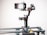 A smartphone is mounted on a motorized gimbal which is attached to a slider. The setup is designed for filming steady video shots. The gimbal has a sleek black design with red accents and features multiple control buttons and adjustment knobs. The background is plain and well-lit, drawing focus to the equipment.