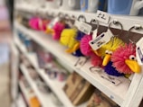 A row of colorful, fluffy keychains shaped like birds hangs on a white shelf. Each keychain has a cartoon-like face with big eyes and a bright yellow beak. They are priced at £2 as indicated by a small sign above them. The shelves appear to be in a gift shop, with other mugs and items slightly blurred in the background.