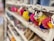 A row of colorful, fluffy keychains shaped like birds hangs on a white shelf. Each keychain has a cartoon-like face with big eyes and a bright yellow beak. They are priced at £2 as indicated by a small sign above them. The shelves appear to be in a gift shop, with other mugs and items slightly blurred in the background.