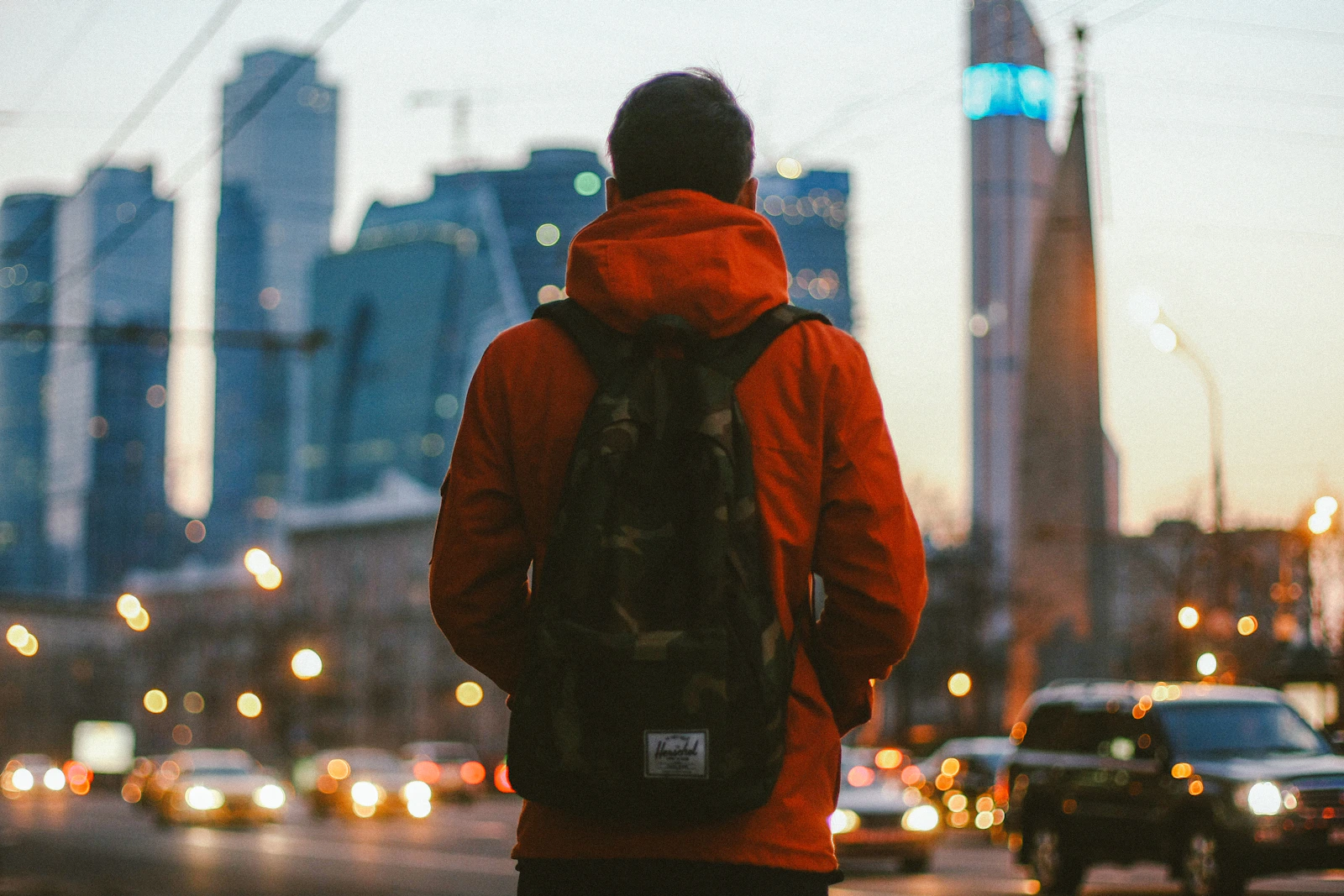 Man in red jacket with backpack, city at dusk