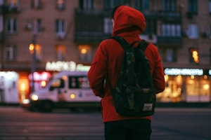 A person wearing a red hoodie and a camouflage backpack stands on a street, facing a blurred cityscape with buildings and illuminated signage. An ambulance or similar vehicle passes by in the background, contributing to an urban nighttime setting.