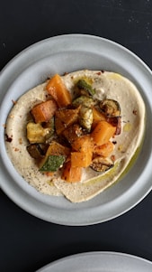 A plate featuring a creamy spread, likely hummus, topped with roasted vegetables including zucchini and sweet potatoes. The dish is garnished with herbs and a drizzle of olive oil, creating an appetizing and colorful presentation.