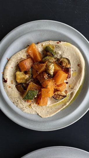 A plate featuring a creamy spread, likely hummus, topped with roasted vegetables including zucchini and sweet potatoes. The dish is garnished with herbs and a drizzle of olive oil, creating an appetizing and colorful presentation.