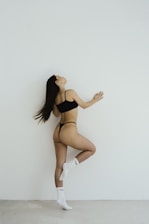 A woman in black underwear and white socks with a swoosh logo poses gracefully against a white wall. Her posture is dynamic, showcasing elegance and poise.