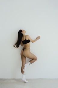 A woman in black underwear and white socks with a swoosh logo poses gracefully against a white wall. Her posture is dynamic, showcasing elegance and poise.
