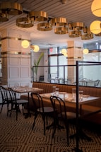 An elegantly designed restaurant interior featuring a contemporary ceiling with circular gold fixtures and round light globes. A row of tables is set with glasses and napkins, accompanied by brown cushioned benches and black wooden chairs. The space is enhanced by patterned flooring and large windows providing natural light.