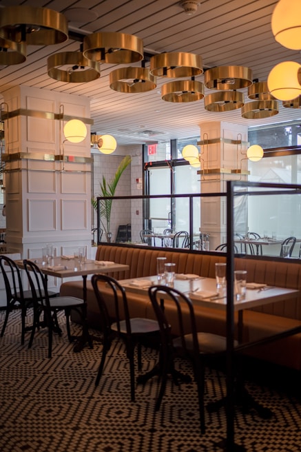 An elegantly designed restaurant interior featuring a contemporary ceiling with circular gold fixtures and round light globes. A row of tables is set with glasses and napkins, accompanied by brown cushioned benches and black wooden chairs. The space is enhanced by patterned flooring and large windows providing natural light.