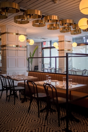An elegantly designed restaurant interior featuring a contemporary ceiling with circular gold fixtures and round light globes. A row of tables is set with glasses and napkins, accompanied by brown cushioned benches and black wooden chairs. The space is enhanced by patterned flooring and large windows providing natural light.