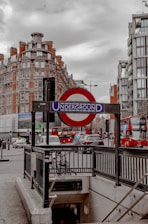 A London Underground entrance is surrounded by urban architecture including brick and glass buildings under a cloudy sky. The iconic red and blue Underground sign is prominently displayed, with double-decker buses and cars visible on the street nearby, indicating a typical busy city scene.