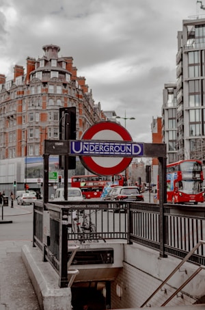A London Underground entrance is surrounded by urban architecture including brick and glass buildings under a cloudy sky. The iconic red and blue Underground sign is prominently displayed, with double-decker buses and cars visible on the street nearby, indicating a typical busy city scene.