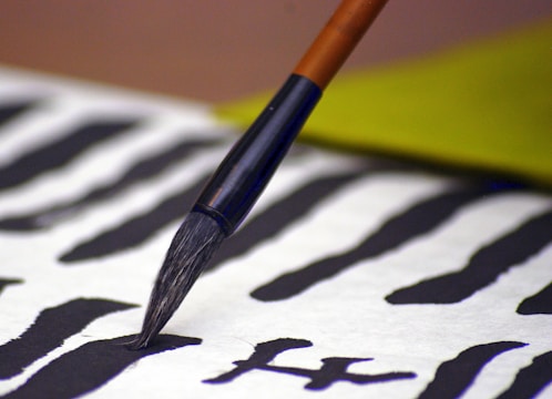 A close-up of a calligraphy brush applying black ink on a paper with abstract black ink patterns. The brush is steady, and the paper shows multiple brush strokes with varying thicknesses. A blurred yellowish-green object is seen in the background, adding a soft color contrast.