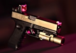 A handgun with a modern design featuring a two-tone finish, with the top part in metallic beige and the lower part in black. It is equipped with a tactical light attachment on the front and a red dot sight on top. The background is blurred and dark, with a reddish hue accentuating the scene.