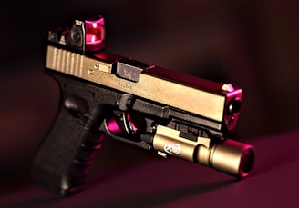 A handgun with a modern design featuring a two-tone finish, with the top part in metallic beige and the lower part in black. It is equipped with a tactical light attachment on the front and a red dot sight on top. The background is blurred and dark, with a reddish hue accentuating the scene.