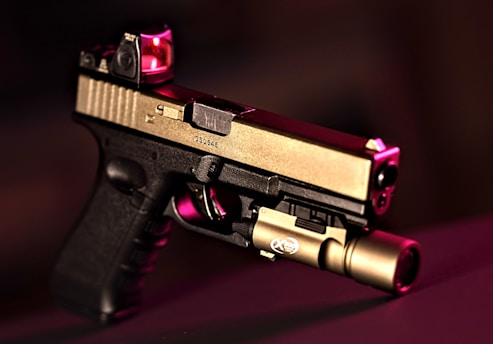 A handgun with a modern design featuring a two-tone finish, with the top part in metallic beige and the lower part in black. It is equipped with a tactical light attachment on the front and a red dot sight on top. The background is blurred and dark, with a reddish hue accentuating the scene.