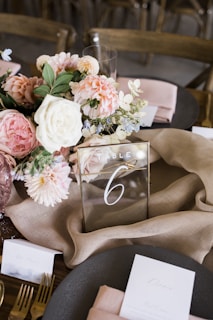 A beautifully arranged table is adorned with a centerpiece of pink and white flowers, including roses and dahlias. A transparent acrylic table number with the number 6 is prominently placed. Surrounding the centerpiece are blush pink napkins neatly folded on dark grey plates, along with gold cutlery. Elegant place cards are positioned beside each setting.