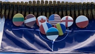 A series of metallic bullets are aligned together, creating an imposing backdrop. Below the bullets, several round badges display flags from different countries, including the United States, Canada, and others, arranged neatly on a NATO flag.