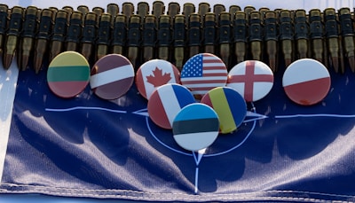 A series of metallic bullets are aligned together, creating an imposing backdrop. Below the bullets, several round badges display flags from different countries, including the United States, Canada, and others, arranged neatly on a NATO flag.