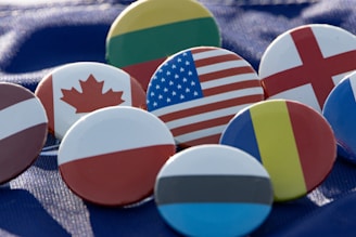 A collection of round pins displaying various national flags, arranged on a blue fabric background. Each pin prominently features the distinct pattern and colors of a different country's flag, such as the United States, Canada, and several European nations.