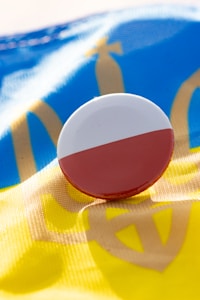 A round pin featuring the Polish flag colors, red and white, is placed on top of a fabric with the Ukrainian flag's blue and yellow colors and a golden trident symbol.