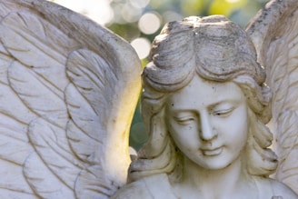 A stone sculpture of an angel with detailed feathered wings. The facial expression is serene, with half-closed eyes and a gentle smile. The soft lighting creates a calm and peaceful atmosphere, highlighting the texture of the stone.