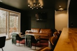 A cozy living room with a modern aesthetic features a brown leather sofa and two black chairs draped with green blankets. A minimalist chandelier hangs from the dark ceiling, providing warm lighting. The room opens onto a wooden deck visible through large glass sliding doors, and a television is mounted on the wall.
