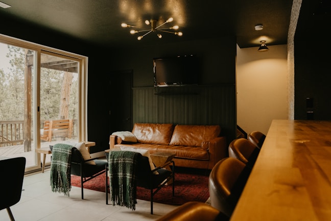 A cozy living room with a modern aesthetic features a brown leather sofa and two black chairs draped with green blankets. A minimalist chandelier hangs from the dark ceiling, providing warm lighting. The room opens onto a wooden deck visible through large glass sliding doors, and a television is mounted on the wall.