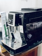 A black DeLonghi cappuccino machine is placed on a countertop. The machine features a control panel with several buttons and a display. A bag of coffee is slightly visible in the background.