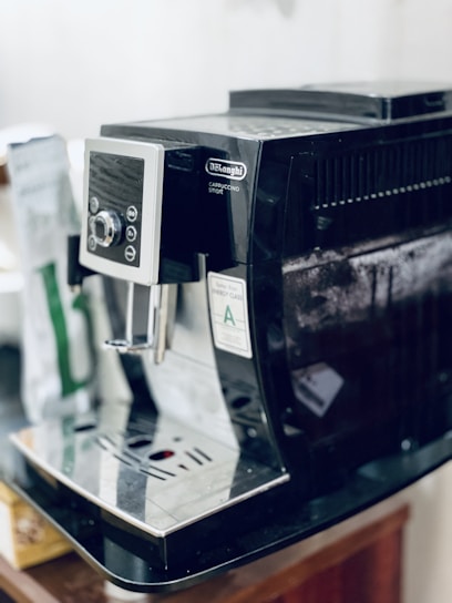 A black DeLonghi cappuccino machine is placed on a countertop. The machine features a control panel with several buttons and a display. A bag of coffee is slightly visible in the background.