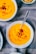 Bright orange creamy soup served in white bowls, topped with croutons and a sprinkle of black pepper, placed on a textured gray cloth.