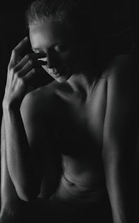 A black and white photograph depicting a person in a contemplative pose, partially illuminated by light that highlights the contours of their face and torso. The individual rests one hand against their face, with an expression that conveys introspection and calmness.