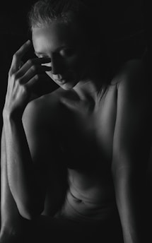 A black and white photograph depicting a person in a contemplative pose, partially illuminated by light that highlights the contours of their face and torso. The individual rests one hand against their face, with an expression that conveys introspection and calmness.